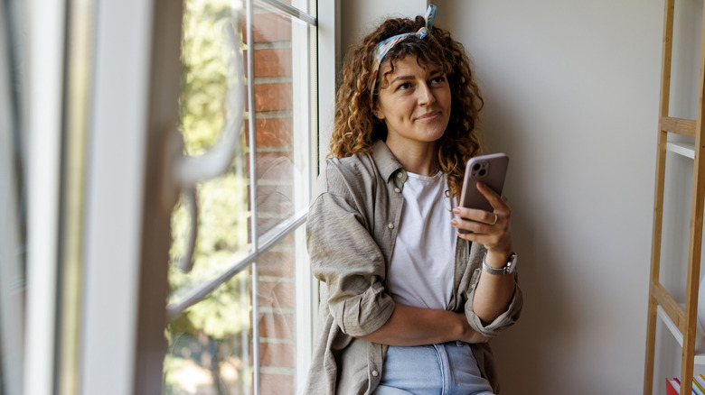 A woman sits by a multipane window and thinks while holding her phone
