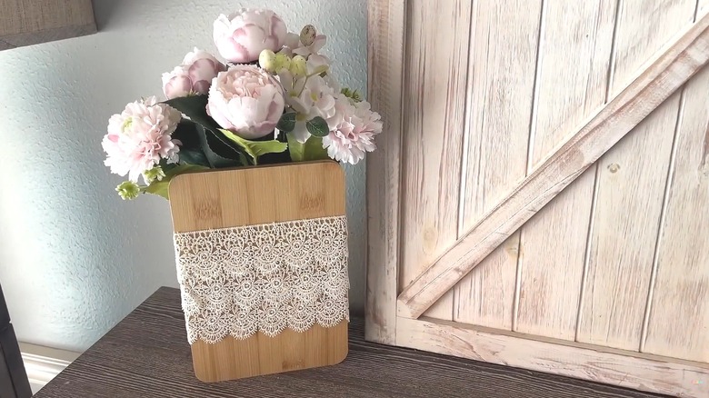 Artificial flowers tucked in a wood cutting board wrapped with lace beside a wood crate in a chic farmhouse room