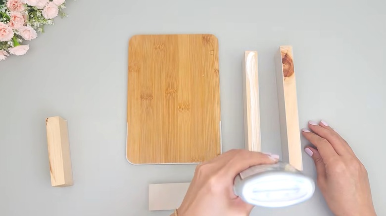 Woman DIYing Dollar Tree cutting board on gray surface with wood craft pieces and pink flowers