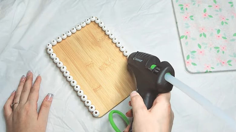 Woman with manicure hot-gluing wood beads to Dollar Tree cutting board on white tarp