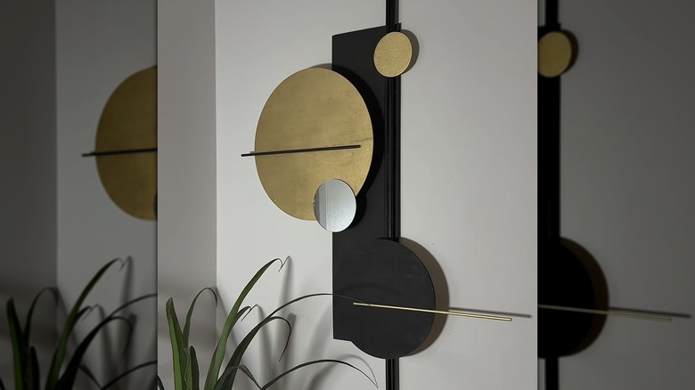 A piece of abstract wall art in black and gold hangs on a gray wall next to a plant