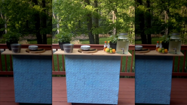 A blue DIY outdoor bar with drinks and accessories on a deck in a backyard with green trees