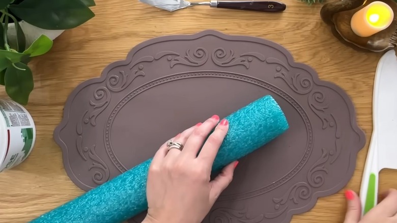 creator holding pool noodle on a brown place mat while reaching for a plastic serrated knife