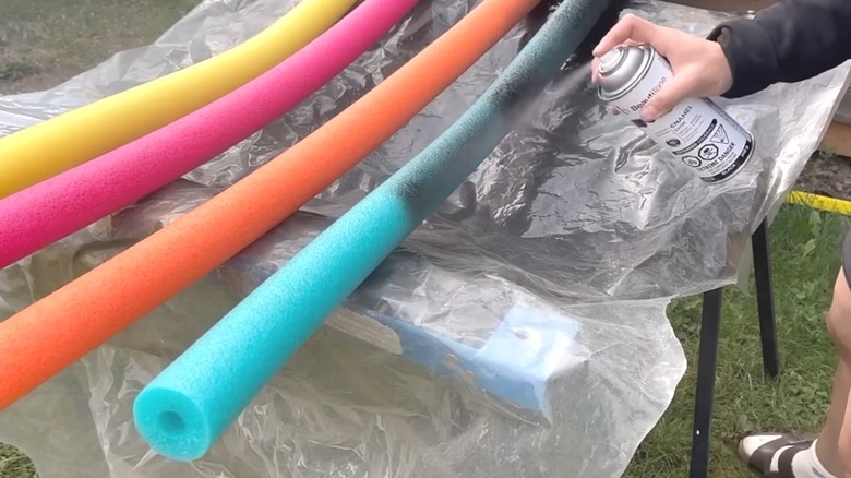 Person spray-painting colorful pool noodles black outdoors on tarp on grass