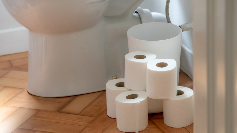Stacks of toilet paper on the floor right next to a white toilet and white trash can