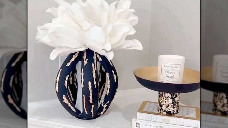 An abstract black and gold vase filled with a large white flower sits on a mantle next to a pile of books and decorative bowl