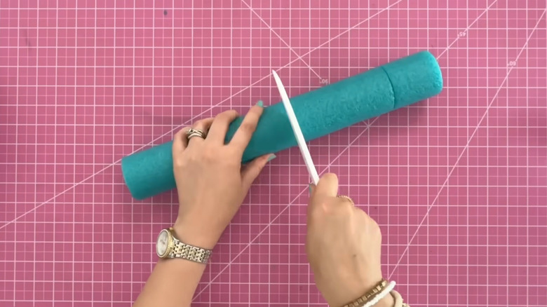 A person cuts into a blue pool noodle on a pink grid surface
