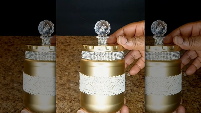 Creator holding a pickle jar painted gold and decorated with lace, gemstone trim, and a bottle stopper