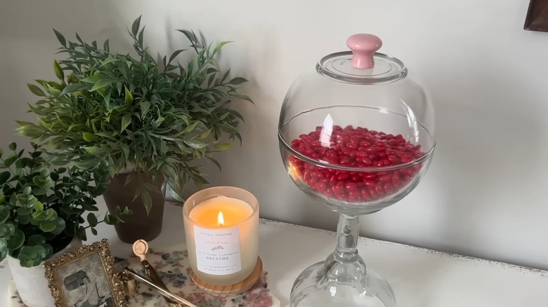 A glass candy dish next to a lit candle, small photo frame, and potted plants