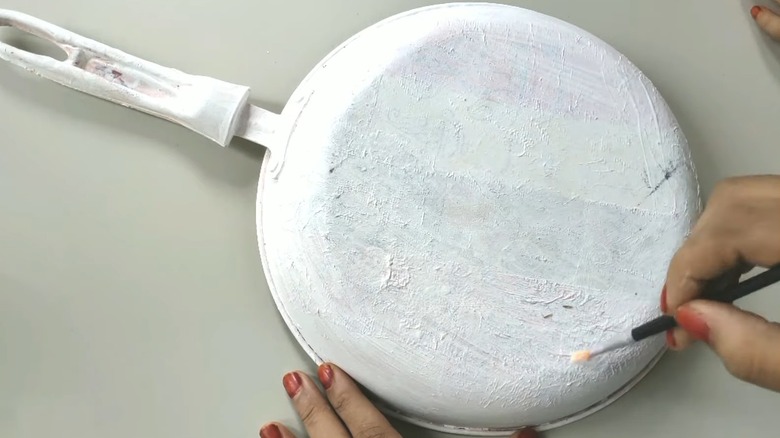 A person with red nails painting old cooking pan white on white table surface with paintbrush