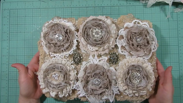 Creator holds up an altered muffin tin featuring fabric flowers, doilies, rhinestones, and other embellishments