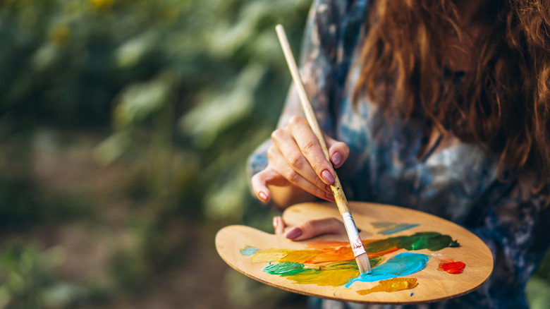 A woman dips her brush into a paint palette in a warm outdoor setting