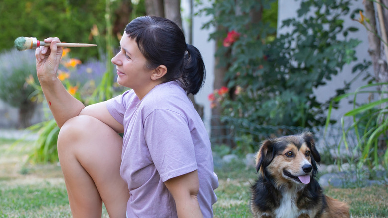 A woman holds a paintbrush while sitting with her dog outside