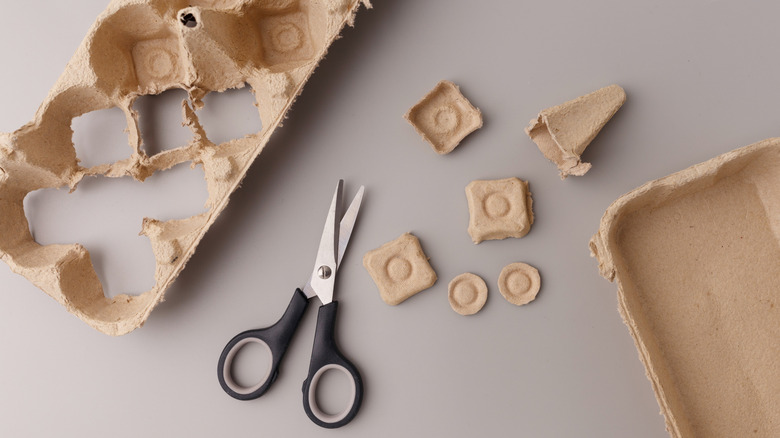 A pair of scissors next to cut up parts of an egg carton on a table.
