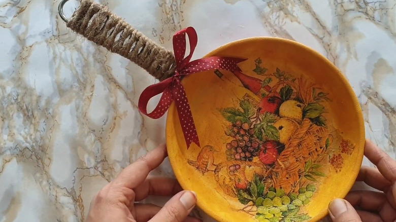 A frying pan with a decoupaged image of a fruit basket, painted yellow, with twine and ribbon on the handle