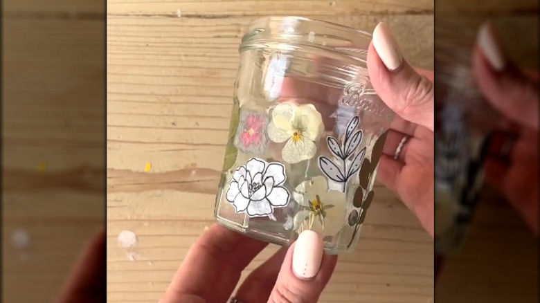 Manicured hands holding a reused jam jar with flower prints and pressed flowers glued around the exterior