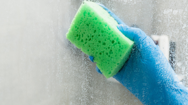 gloved hand with sponge wiping stained shower glass