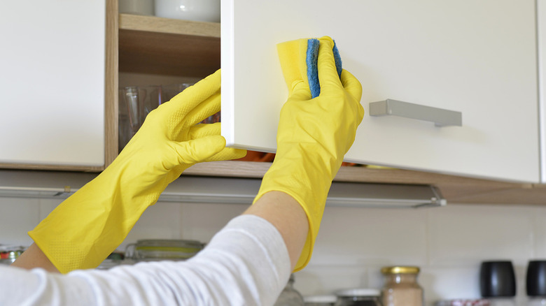 Yellow gloved hands clean a white kitchen cabinet using a sponge.