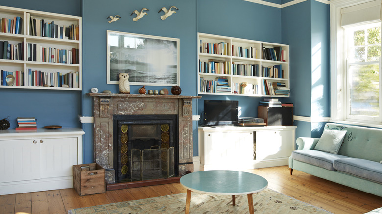 A living room is painted a dark turquoise. There are built-ins on either side of the fireplace and a sofa under the large window.