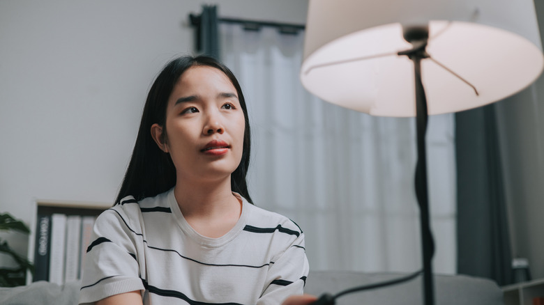 A woman looking thoughtfully at a beige table lamp in a bedroom