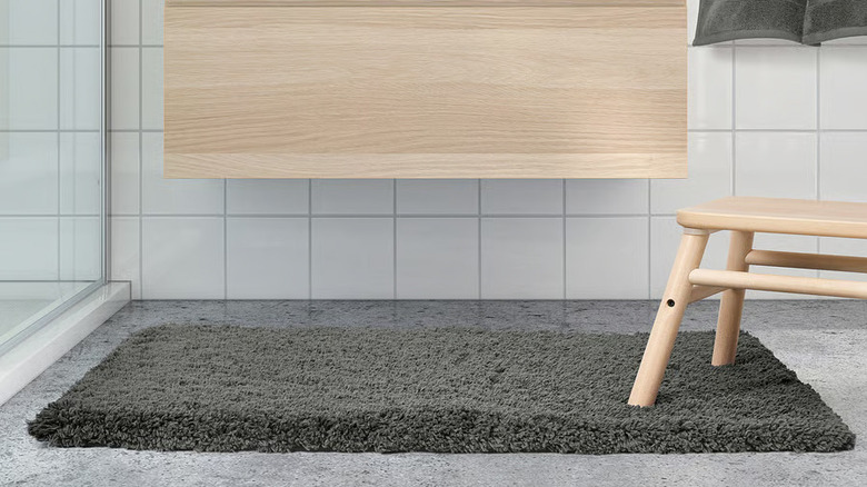A thick gray bathmat sits on a gray floor below a light wood wall-mounted bathroom cabinet