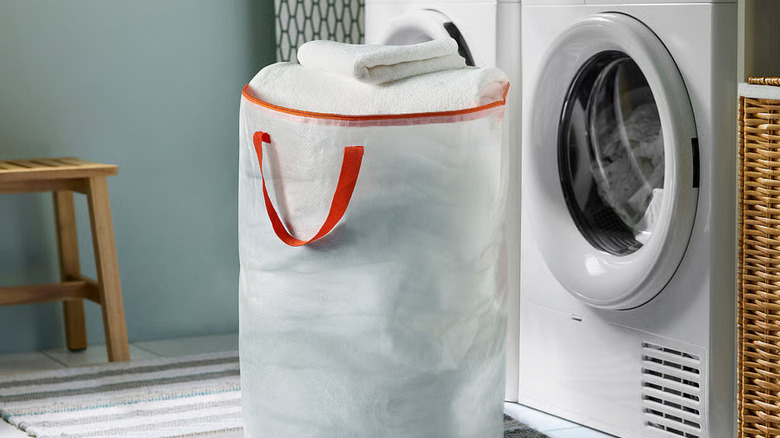 An upright laundry bag with orange handles sits in front of a washing machine