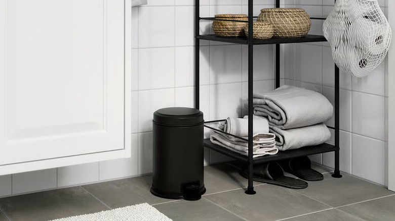 A small black trashcan sits on a bathroom floor between a shelving unit and a white cabinet