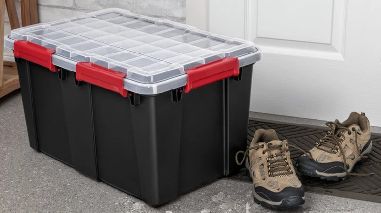 A black storage tote with a clear lid and red latches sits on a concrete floor next to a pair of work boots