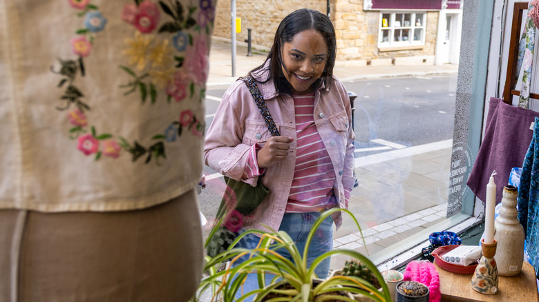 A woman looks inside a thrift store window, with candleholders, vases, plants, and textiles in the shop