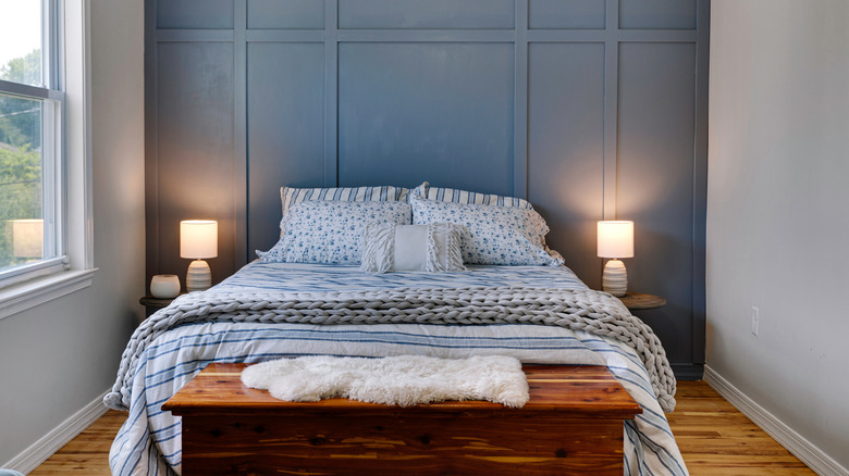 A small bedroom with wood floor, wood trunk, side tables, bed with blue bedding and a blue accent wall with board and batten