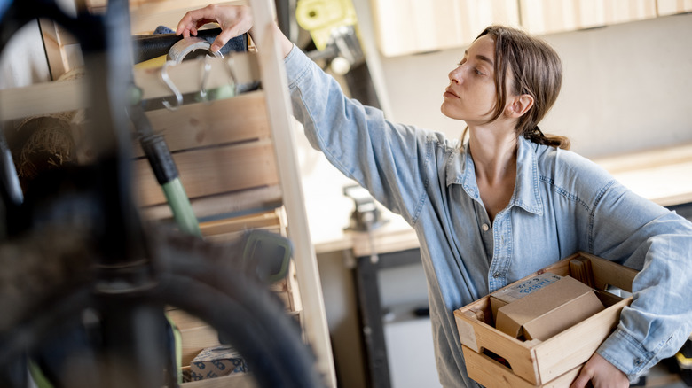 A woman reaching for something on a garage shelf while holding a bin in hand