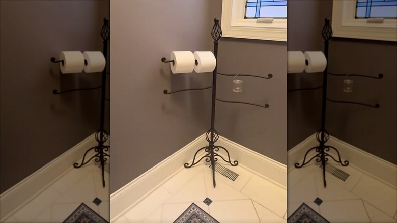 Freestanding iron towel hanger in the corner of a bathroom with two rolls of toilet paper and hanging candle holders on it