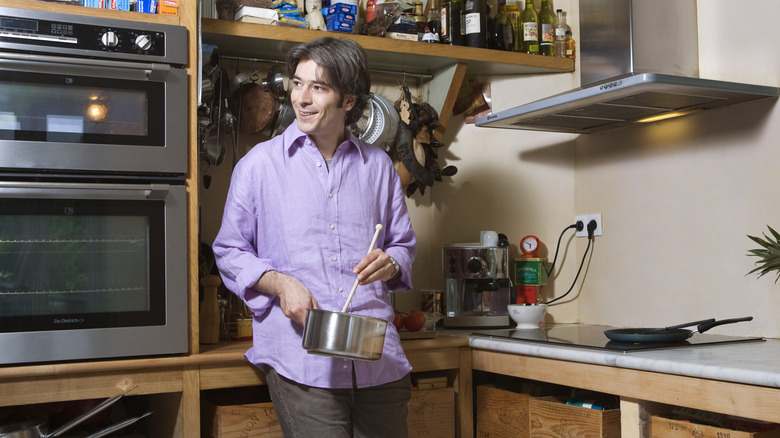 A person in a light purple shirt stirring a pot in a cluttered kitchen