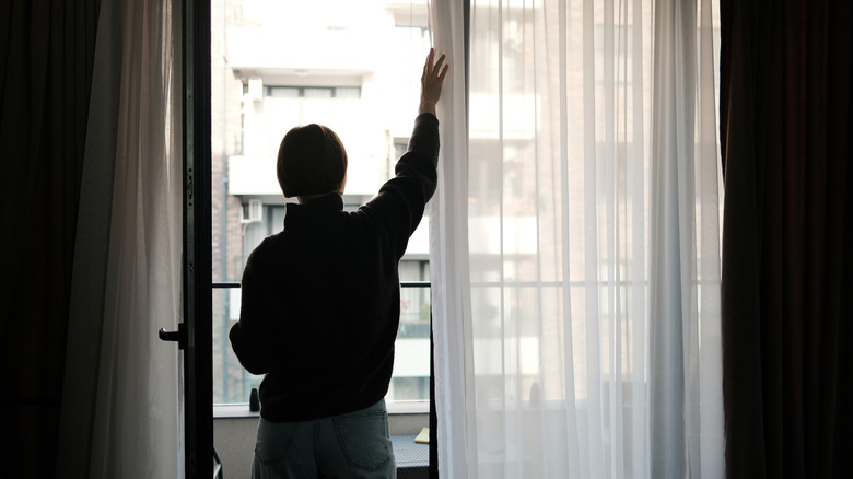 Person pulling back white curtains away from glass balcony door