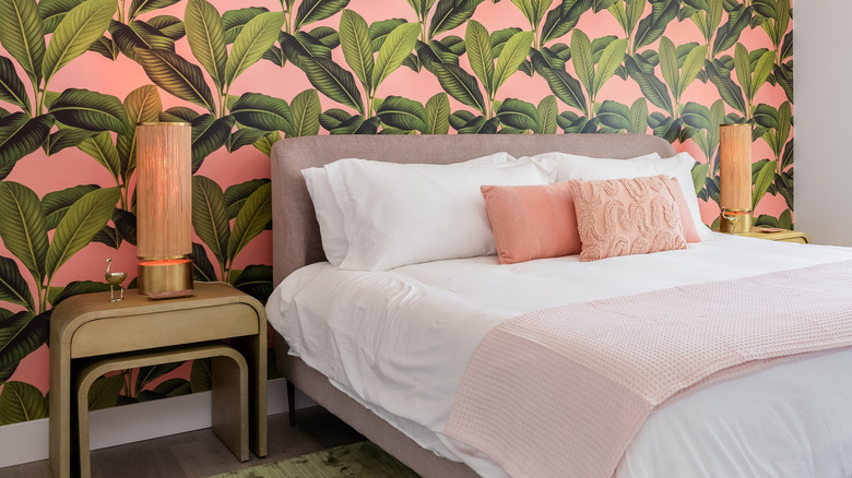 Bedroom with white bed, pink pillows, and pink wallpaper with tropical plant patterning.