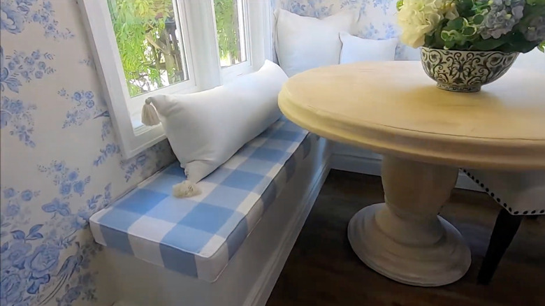 A blue-and-white gingham cushion sits on a white L-shaped banquette in front of a pedestal dining table.