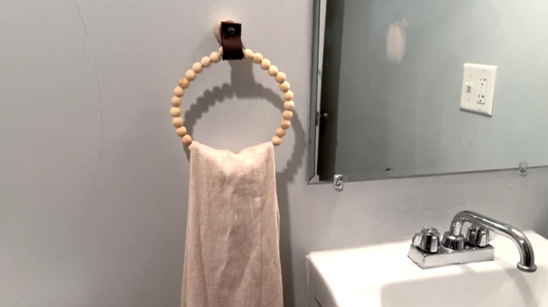 A beige bath towel hanging from a beaded towel ring next to a sink and a mirror