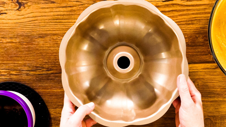 A woman's hands on a bundt pan on a wooden table next to grease and some sort of mix