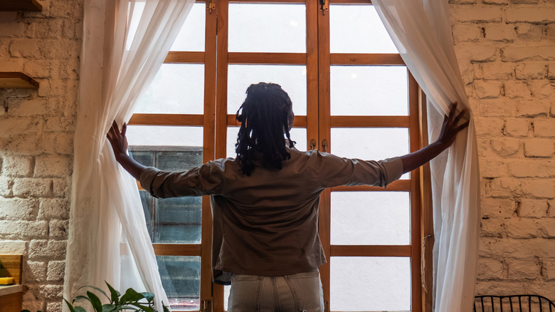 A woman opens plain curtains in a rustic room