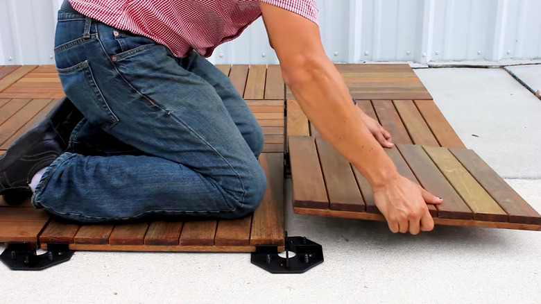 A worker kneels while placing a composite wood deck tile into place over a concrete patio