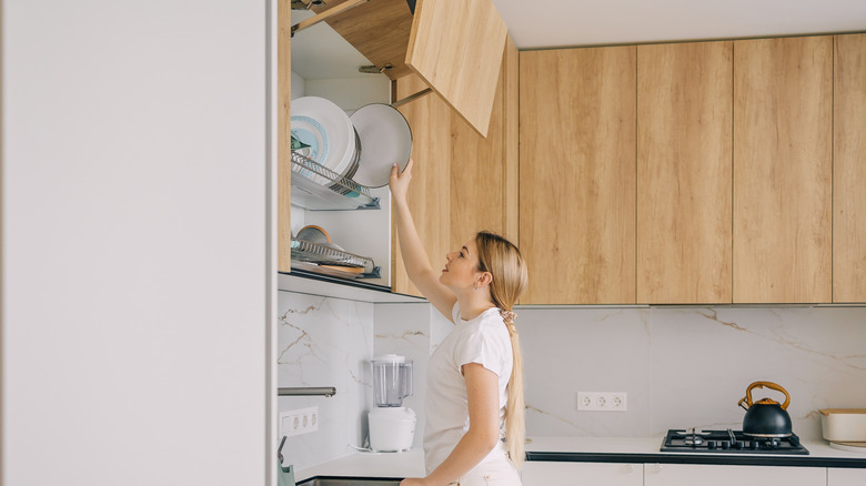 Woman reaching upward to grab plate from overhead kitchen cabinet