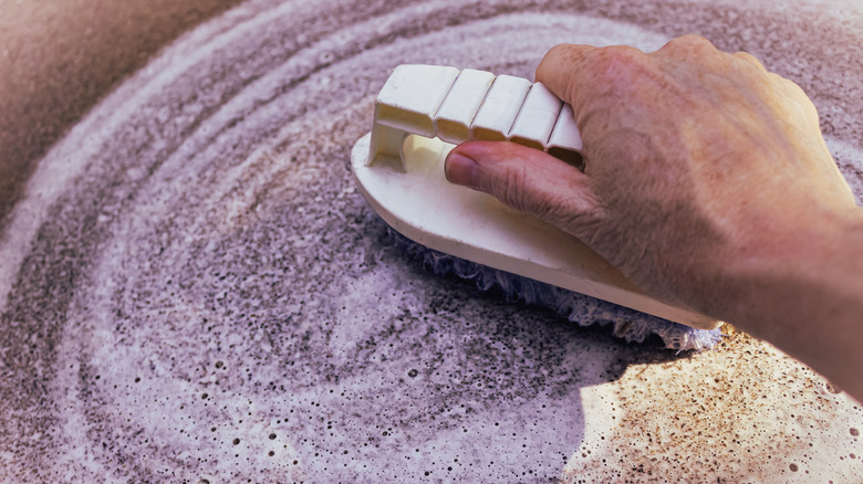 Closeup of cleaning in progress: A hand actively scrubbing a concrete surface with a brush to remove stains with a foam background.