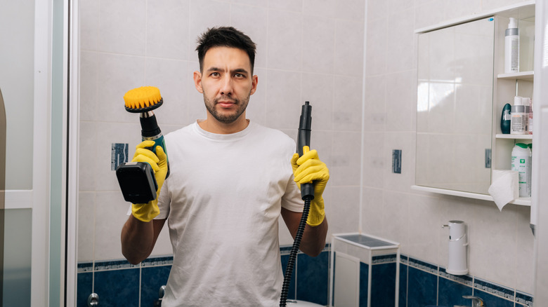 A person holding a steamer and a drill-powered scrub brush in a bathroom.