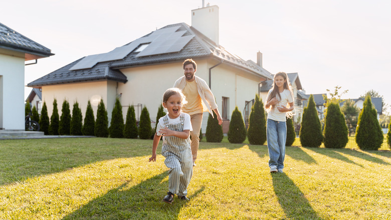 Father playing with his daughters in the yard with solar panels on the roof in the background.