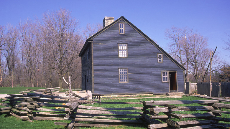 A blue saltbox house with a sloping roof and wood surrounding it like a fence
