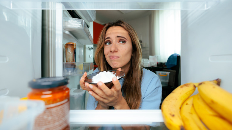 A view of a woman from inside the fridge. She's holding baking soda and making an odd face