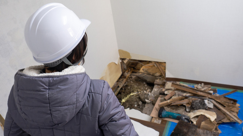 A person wearing a hardhat looks at a rotten floor in the corner of a flooded room