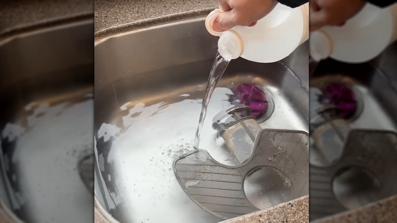 A person adding vinegar to a sink filled with warm water to soak a dishwasher filter,