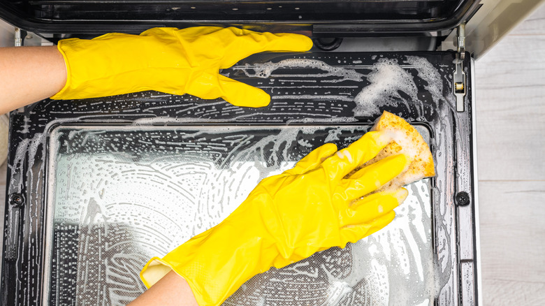 Two yellow-gloved hands cleaning oven door with sponge and soapy solution