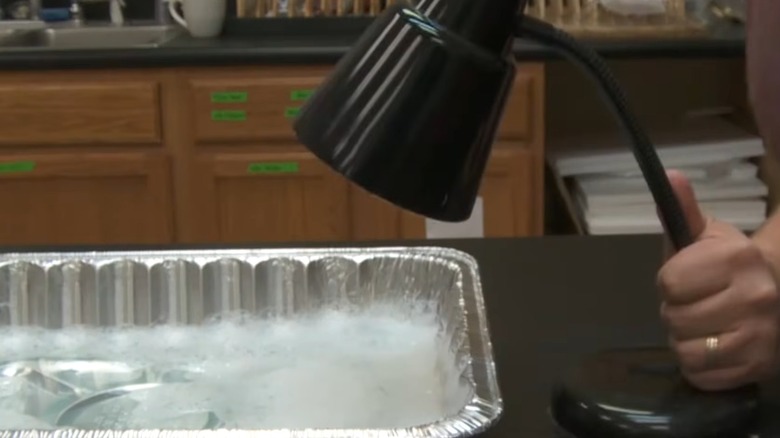 Hand placing a black table lamp over a roasting aluminum pan holding soapy water